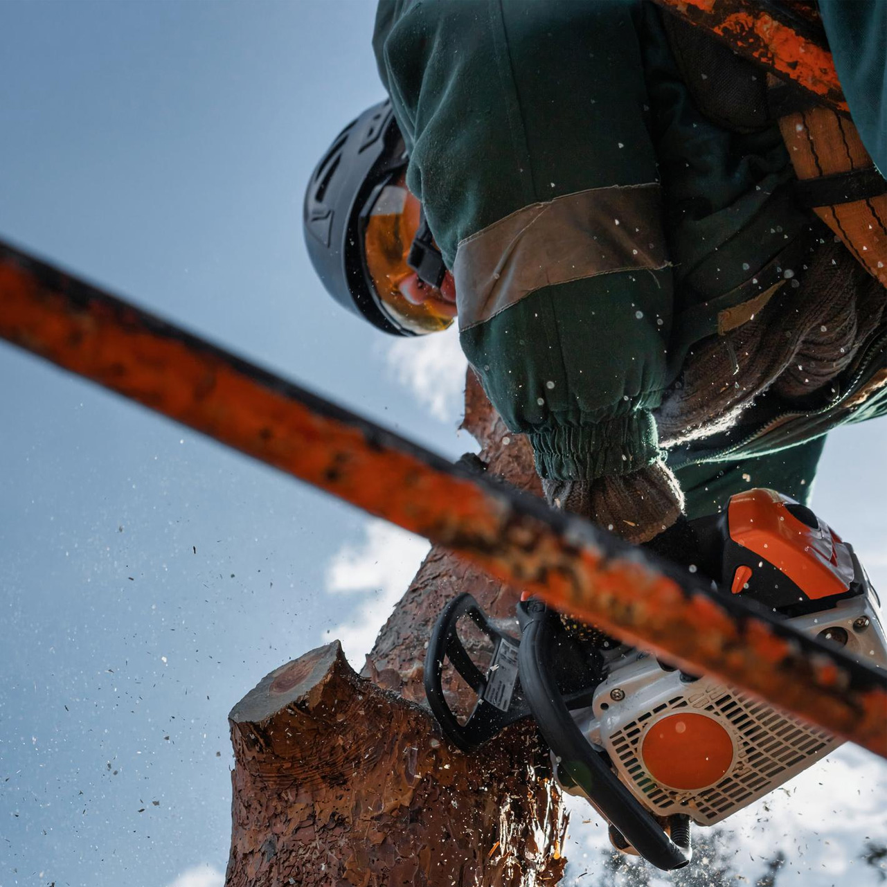 Arborist using a chainsaw to cut a tree branch while secured by safety gear and ropes against a backdrop of blue sky.